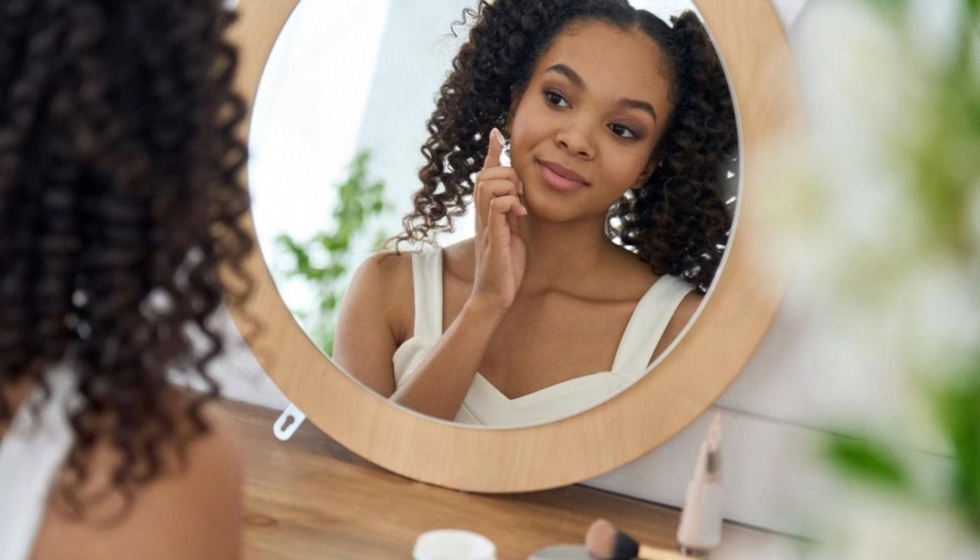 Young woman looking in a round mirror while applying serum to her cheek as part of a daily preventive skincare routine