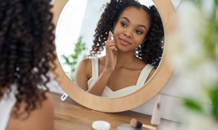 Young woman looking in a round mirror while applying serum to her cheek as part of a daily preventive skincare routine